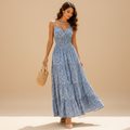 A woman models a light blue floral maxi dress with thin straps, a V-neckline, and a tiered skirt, holding a woven straw handbag against a solid beige studio background.
