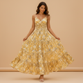 A woman modeling a yellow floral maxi dress with thin spaghetti straps, a V-neckline, and a tiered ruffled skirt, set against a solid beige studio background.